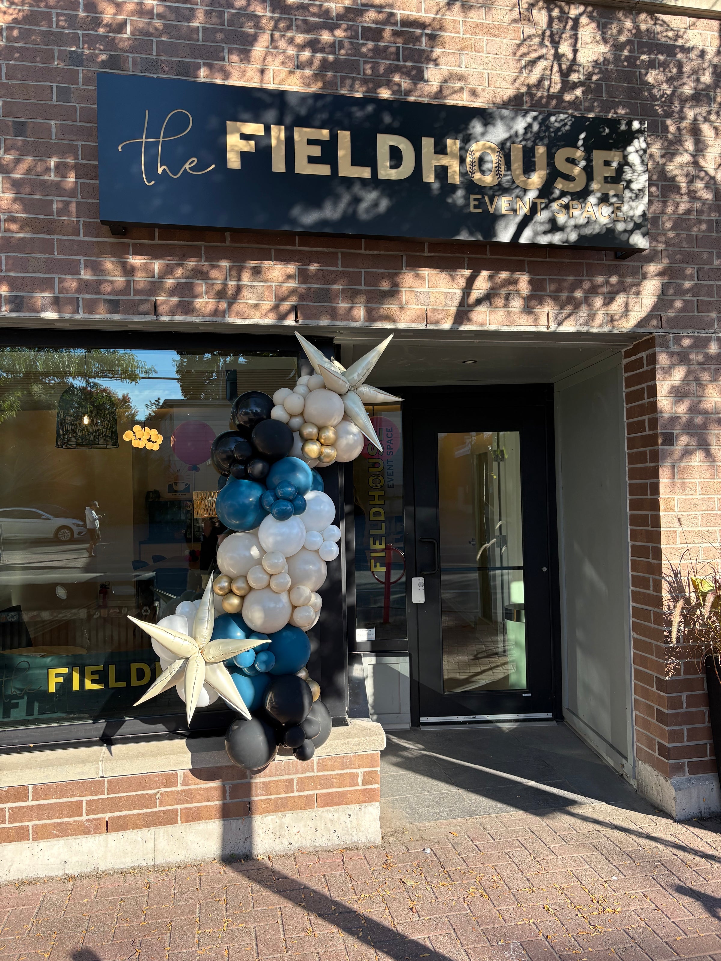 balloon garland in black, deep teal, sand, gold and white colours with starburst balloon accents in beige colour. Installed outdoors at front of a retail shop.