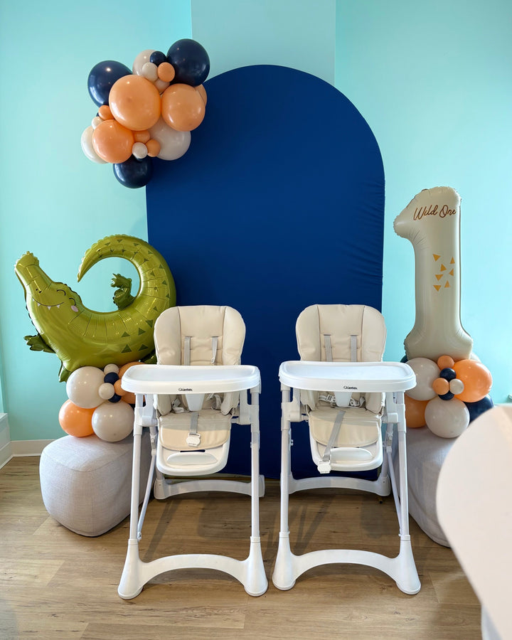 Two high chairs with balloon columns on either side and a small balloon garland mounted on a blue backdrop.
