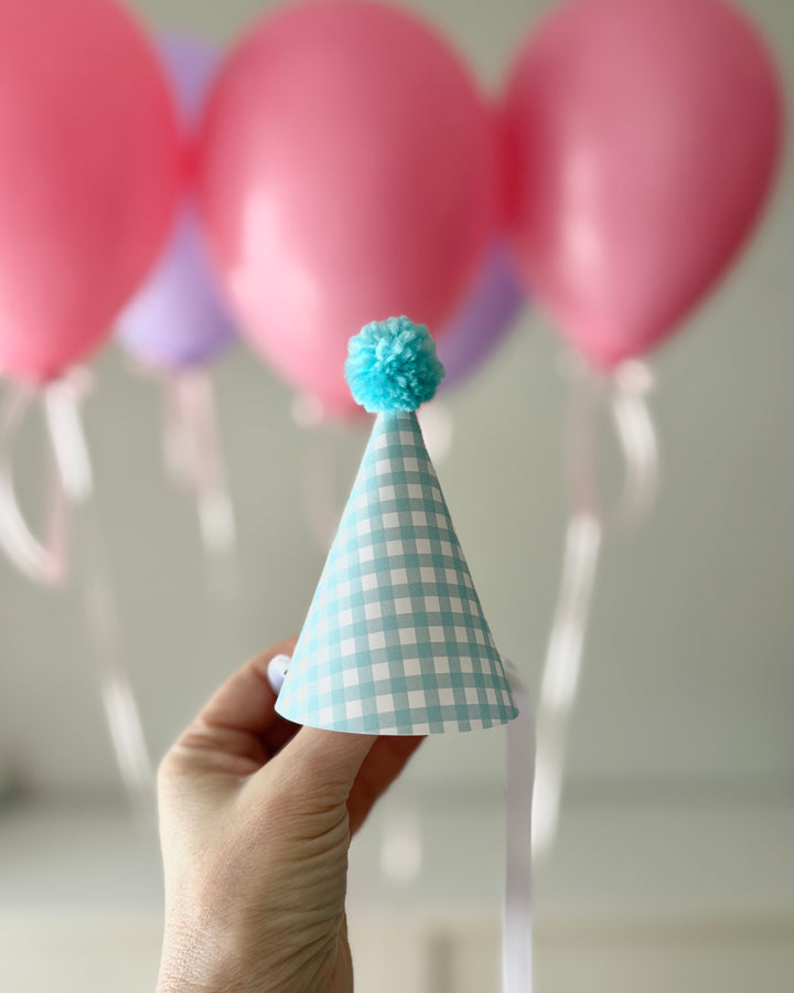 Hand holding a blue mini checkered party hat with a blue pom-pom against balloons.