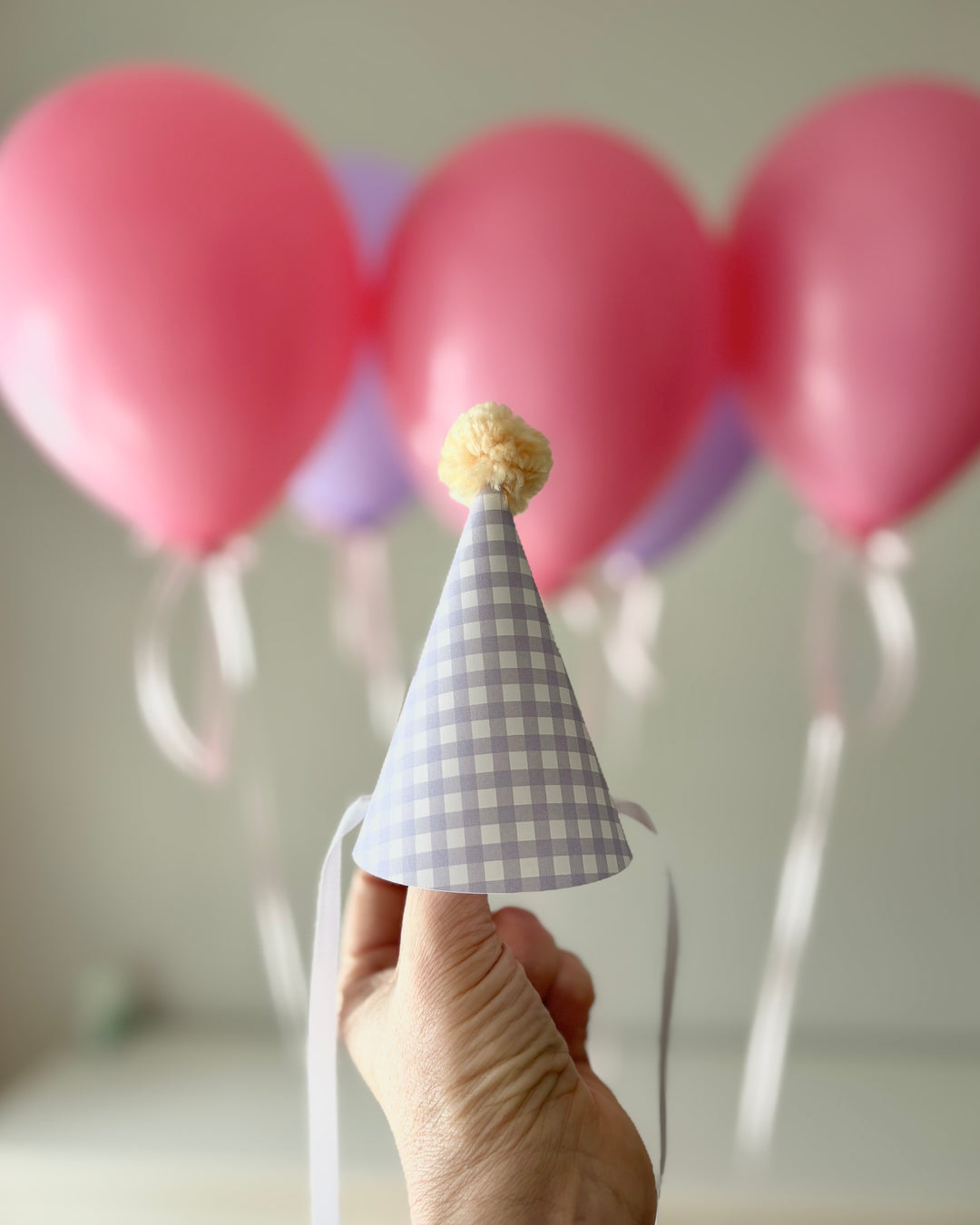 Hand holding a lilac checkered party hat with pink balloons in the background