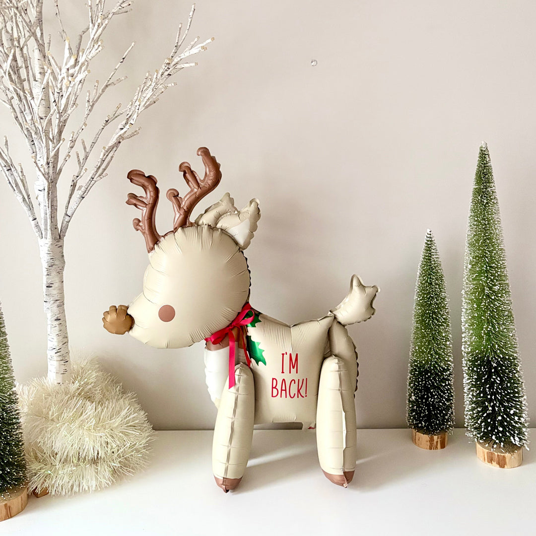 Decorative reindeer balloon with 'I'm Back!' text, surrounded by Christmas trees and branches on a neutral background. Elf  on the shelf arrival balloon