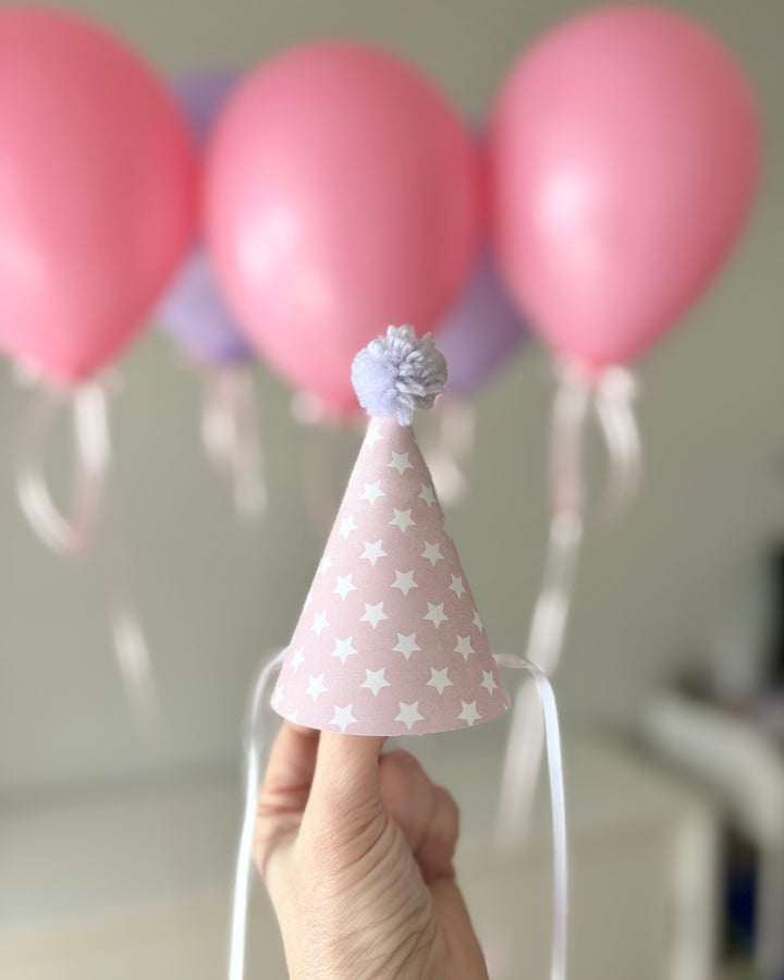 Hand holding a pink mini party hat with star pattern in front of pink balloons