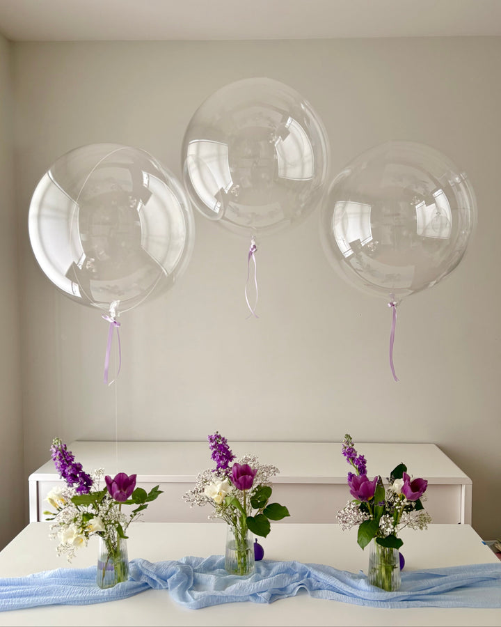 Clear bubble balloons with ribbons on a plain wall above a table with floral arrangements.