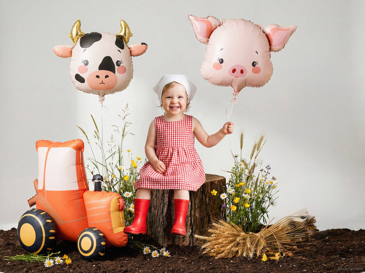 Child in a checkered dress with animal-shaped balloons and a toy tractor on a farm-themed set.