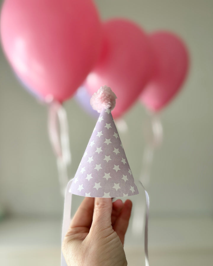 Hand holding a lilac mini party hat with star pattern in front of pink balloons