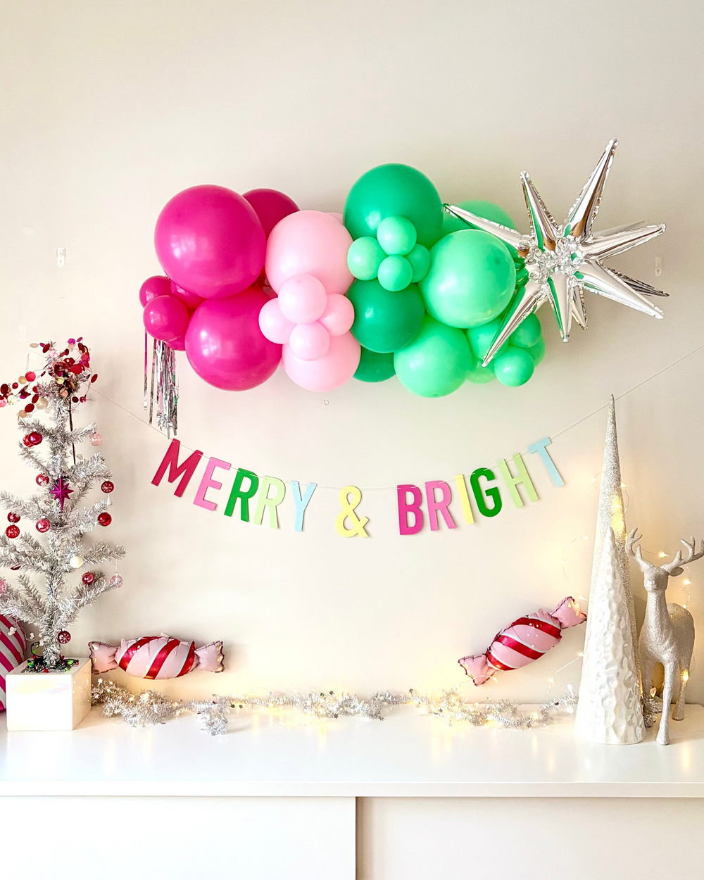 Decorative setup 'Merry & Bright' banner, Christmas-themed ornaments, and a balloon garland with silver accents, on a white wall.