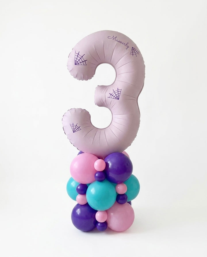 A jumbo pink number 3 balloon in a pale pink-lavender color with purple spiderweb designs, standing upright on a coumn of pink, purple, and teal balloons against a clean white background.