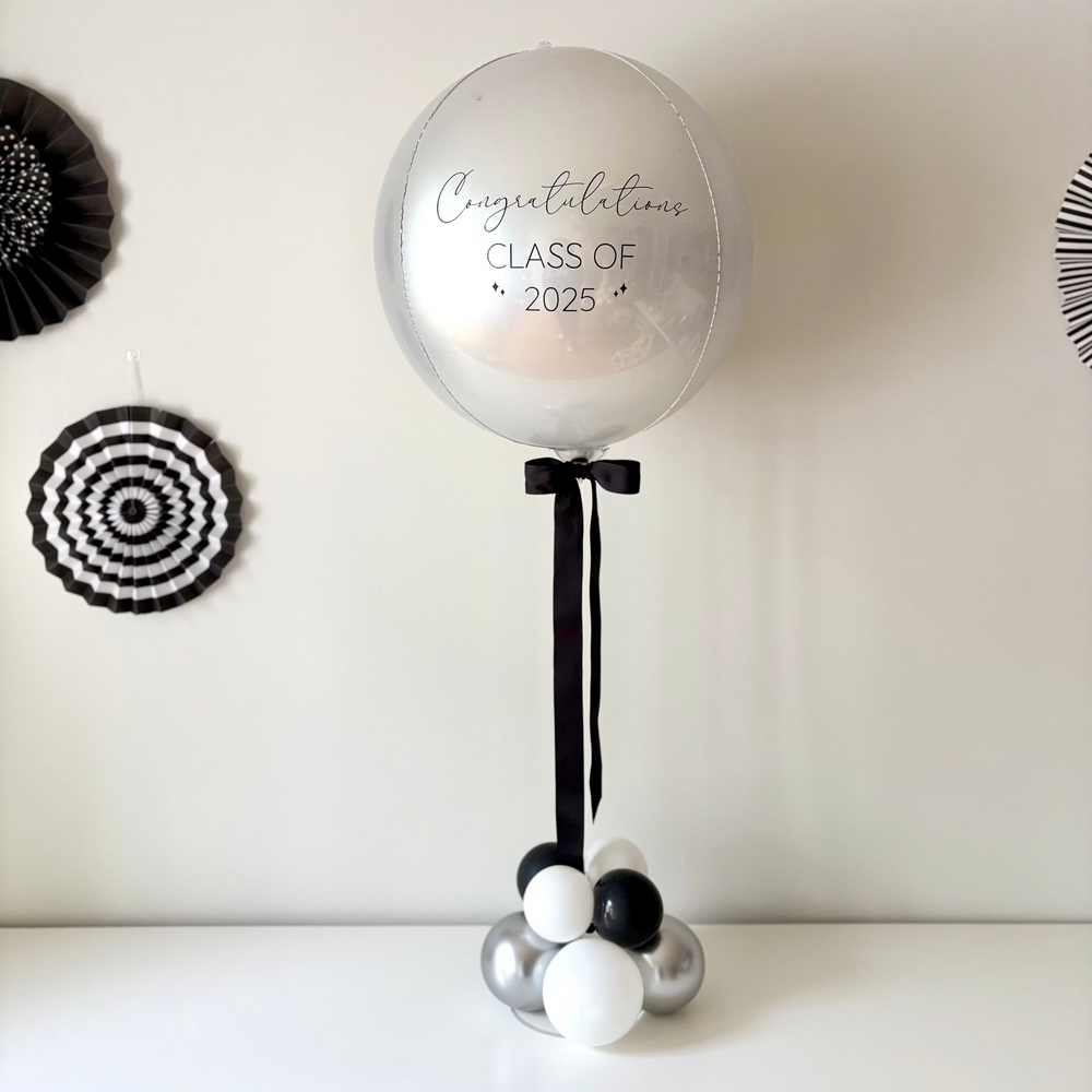 A balloon table centrepiece featuring a large silver balloon with a custom message, mounted on a clear acrylic base, with a smaller black and white balloon and a satin ribbon bow.
