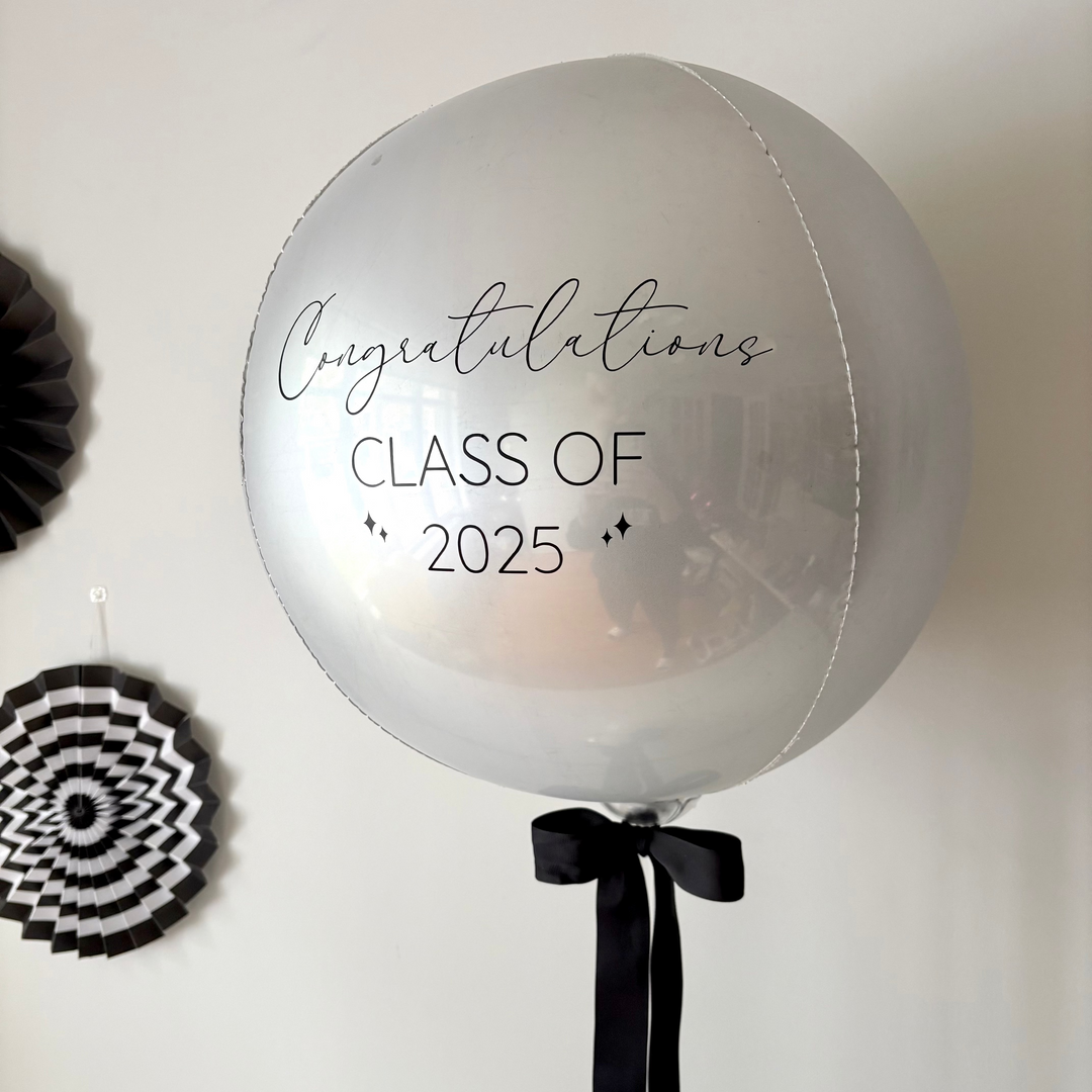 Close up of a balloon table centrepiece featuring a large silver balloon with text "Congratulations Class of 2025" and black bow