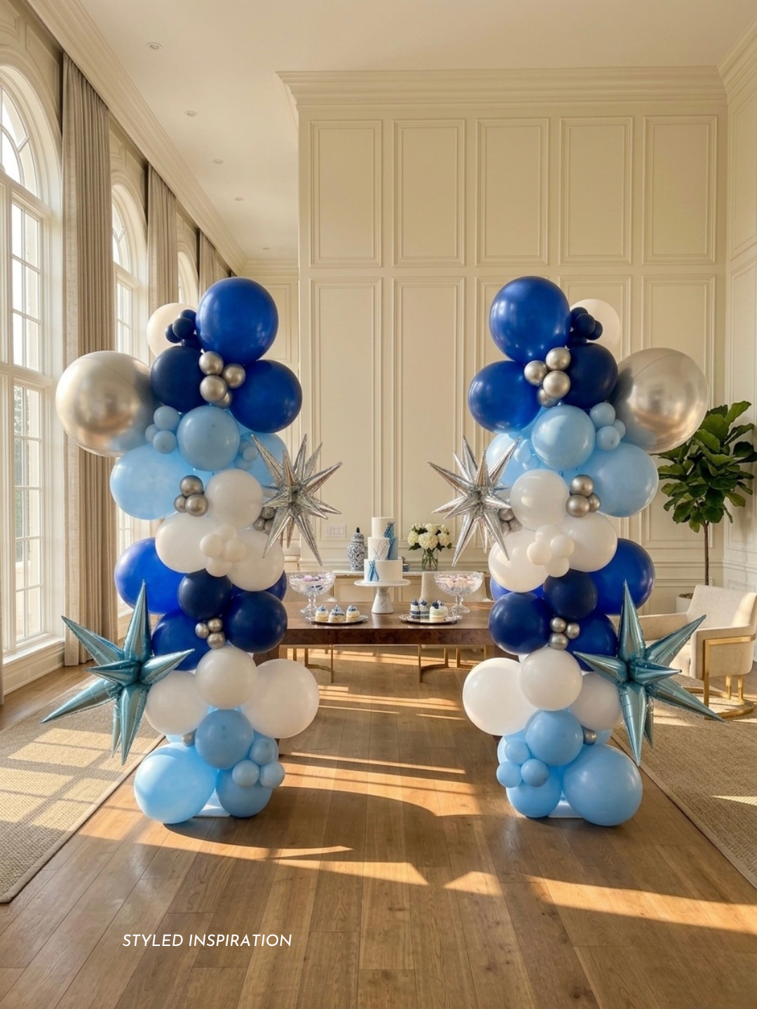 pair of organic balloon column arrangement with metallic star shaped 3D balloons starbursts and orbz (sphere) balloon, styled in a decorated room with tall windows on the left and a table with a cake behind 