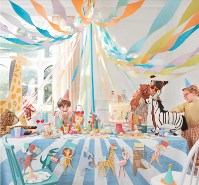 Scene of Animal Parade Party with colourful paper streamers from ceiling, happy children sitting around table decorated with animal parade garland and cake and cute animal cups